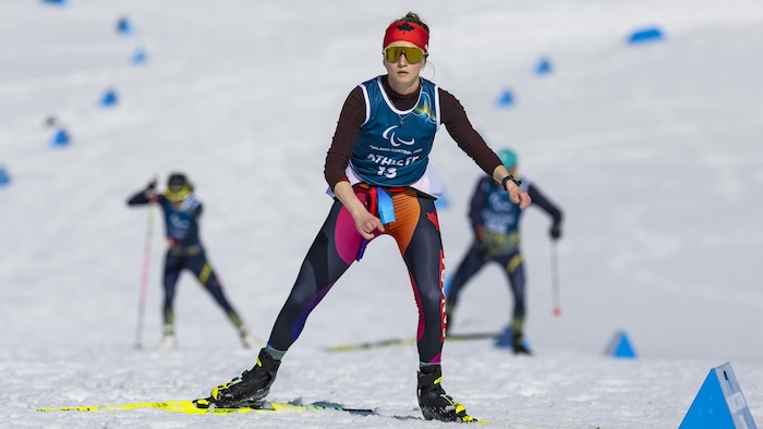Une skieuse de fond skie, sans utiliser de bâtons. Elle porte des lunettes de soleil et un bandeau rouge avec un dessin de feuille d'érable noire.