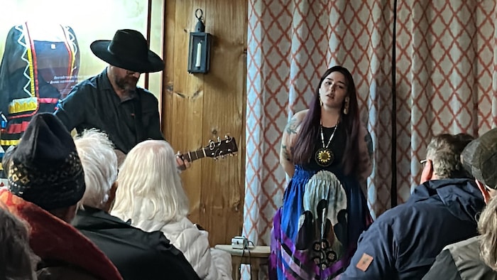 Emma Stevens est debout devant l'audience, les mains dans le dos, un rideau blanc et rouge derrière elle et un guitariste à sa gauche.