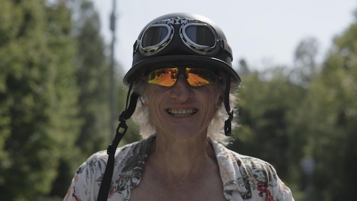 Un homme portant un casque de moto et des lunettes de soleil sourit à l'extérieur.