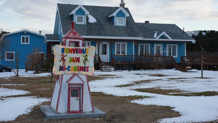 Une maison sur un terrain recouvert partiellement de neige. Au premier plan un phare miniature sur lequel est apposé une pancarte où il est écrit « Bienvenu aux mi-carêmes ».