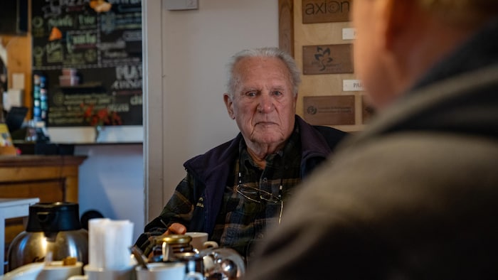 Un aîné regarde droit devant. Il a une tasse à café dans la main gauche.