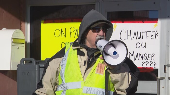 Devant une affiche sur laquelle est écrit «Chauffer ou manger????», un homme portant une veste de sécurité jaune, le capuchon rabattu sur la tête, est debout en public et parle dans un mégaphone.