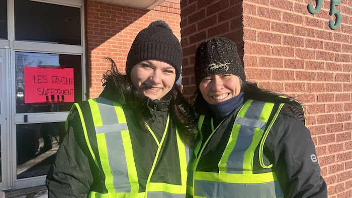 Deux femmes posent ensemble et sourient devant un édifice. Elles portent des tuques et des vestes réfléchissantes jaunes.