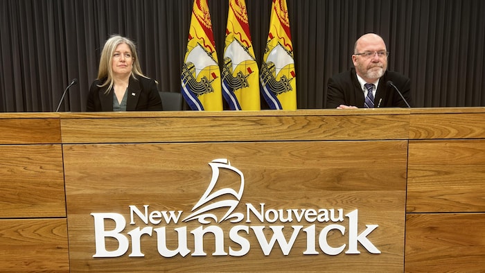 Une femme et un homme assis devant des micros, des drapeaux du Nouveau-Brunswick derrière eux.
