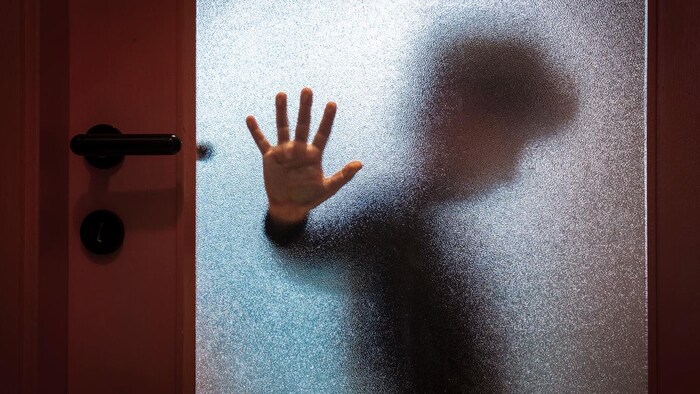 Un enfant derrière une fenêtre givrée pose sa main contre la vitre et regarde au sol.
