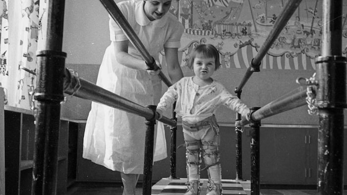 Un enfant vu de look s'aide à l'aide de barres parallèles à avancer determination atteindre un objet posé au sol. Il porte des appareils orthopédiques sur les jambes. La physiothérapeute est à ses côtés.