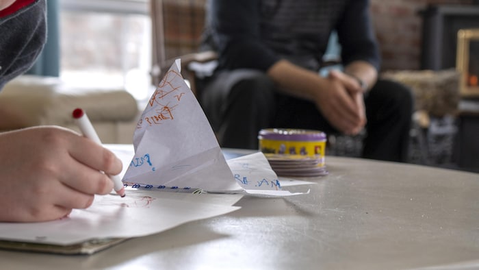 Gros plan sur une main d'enfant qui dessine et un adulte assis sur une chaise en arrière-plan.