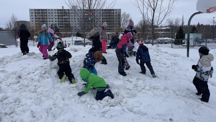 Un groupe d'enfants s'amuse à grimper sur un imposant amas de neige dans une cour d'école.