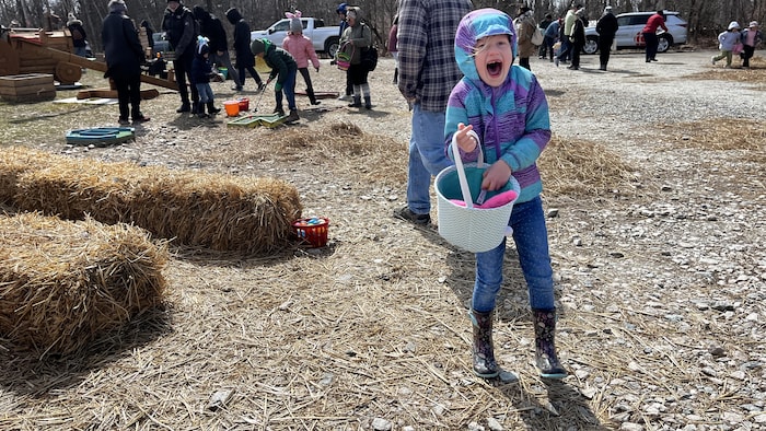 Une petite fille sourit avec une dent manquante, une main dans son panier plein d'œufs de Pâques