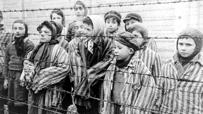 Des enfants ayant survécu au camp de concentration d'Auschwitz, vêtus de vestes de prisonniers pour adultes, se tiennent derrière une clôture barbelée en Pologne.