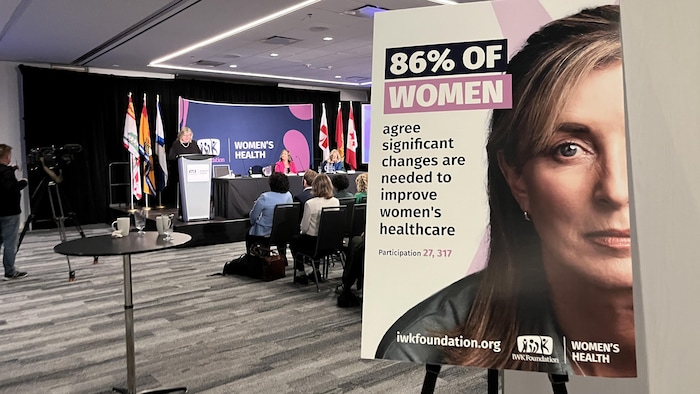 Une affiche montrant le visage d'une femme et le titre « 86 % des femmes » est posée à l'entrée d'une salle de conférence où une femme prononce une allocution devant public.