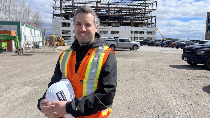 Un homme pose devant un chantier de construction avec un casque dans ses mains.
