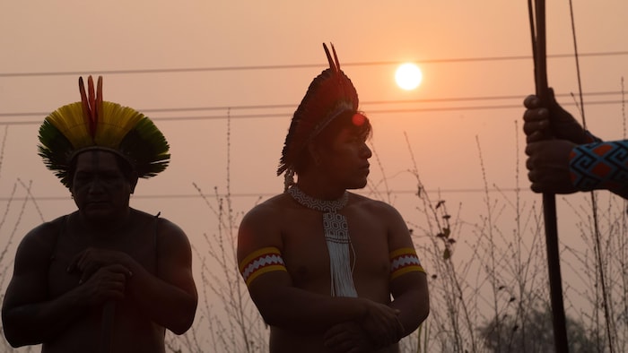 Trois Autochtones de l'Amazonie brésilienne en habits traditionnels se tiennent debout devant un coucher de soleil.