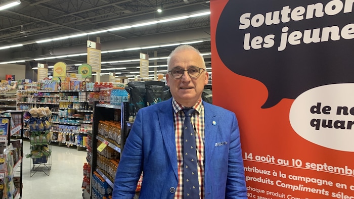 Un propriétaire d'épicerie pose devant une affiche posée devant des allées.