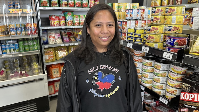 Une femme avec un chandail noir dans une épicerie où plusieurs produits sont visibles.