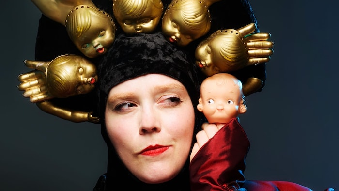 L'artiste Ingrid Hansen porte une coiffe de têtes de poupées et tient une figurine près de son visage.