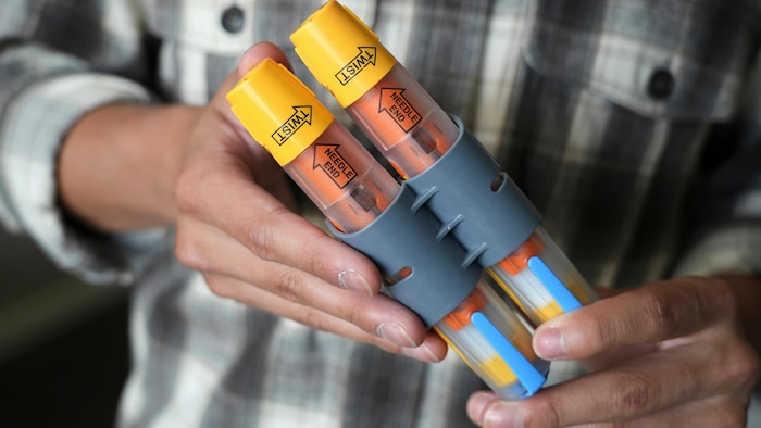 Plan rapproché de deux auto-injecteurs tenus par les deux mains d'un homme portant une chemise à carreaux.