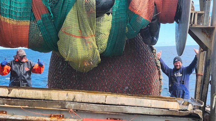 Deux hommes souriants lèvent les pouces de chaque côté d'un énorme filet sur le pont d'un bateau en mer.