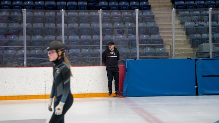 Éric Bédard est debout en patins sur une glace, derrière une patineuse de vitesse.