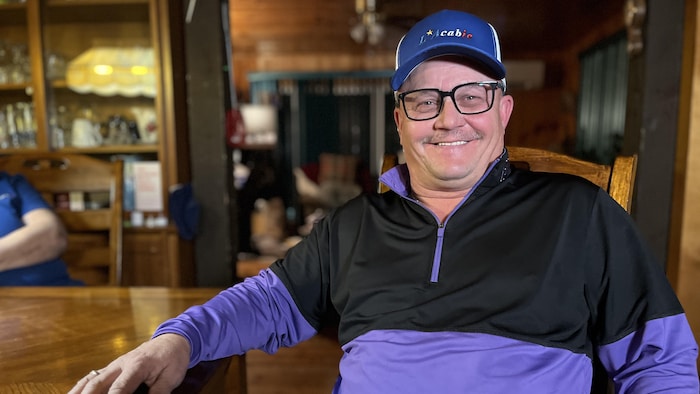 Un homme avec une casquette est assis à une table. Il se tourne vers la caméra et fait un grand sourire.