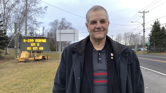 Éric Dubé sourit devant un panneau indiquant la fermeture de la route 299 du kilomètre au kilomètre 60.