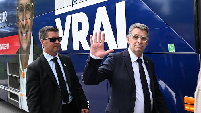 Éric Duhaime salue de la main devant un autobus bleu de campagne, aux côtés d'un agent de sécurité en costume.