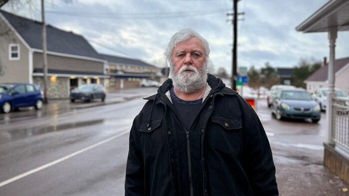Un homme à la barbe blanche hirsute est debout dans une rue mouillée, sous de sombres nuages, et a un air lugubre.