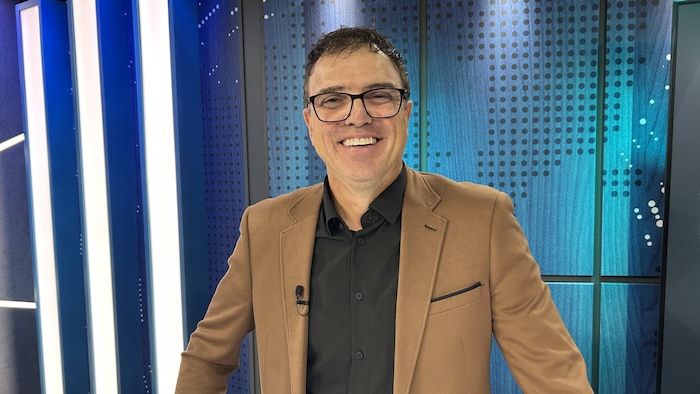 Un homme en complet sourit à la caméra sur un studio de télévision.