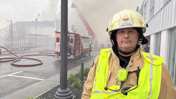 Éric Hébert est vêtu de son casque et de son habit de protection, en plus d'un dossard de sécurité. Il pose dans un nuage de fumée, sur le bord de la rue, où des camions de pompiers sont stationnés. Des boyaux d'incendie sont dans la rue.