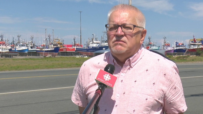 Un homme debout en bordure de la route en été. On voit un quai et des bateaux derrière lui.