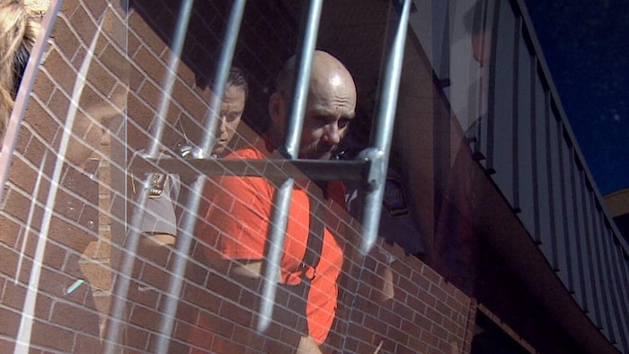 Le reflet d'un homme au crâne rasé, vêtu d'un t-shirt orange, derrière des barreaux en métal et entouré de deux policiers en uniforme.