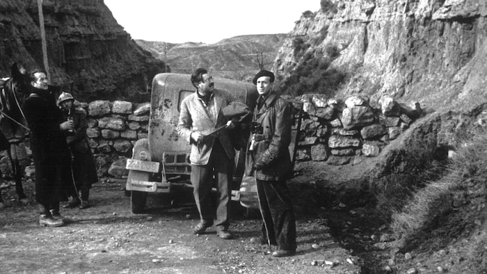 Quatre hommes sont sur un petit chemin, près d'une voiture et d'un cheval. Trois d'entre eux regardent quelque chose qui semble se situer dans une montagne.