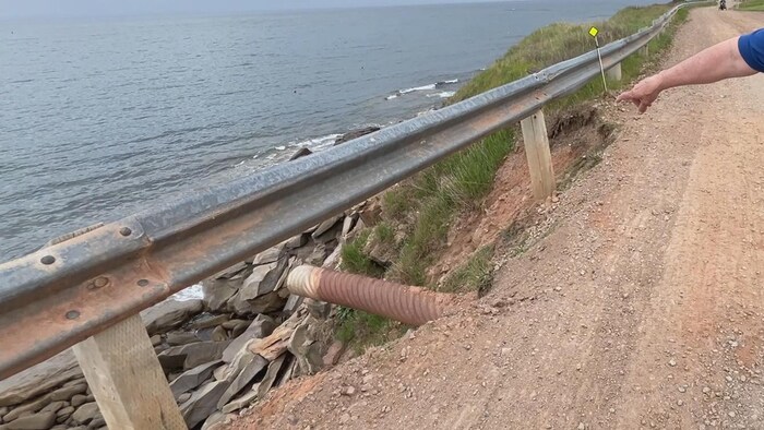 Un homme debout sur un chemin de terre au bord de l'eau pointe un tuyau en métal qui sort d'en dessous de la route et s'étend au-dessus du vide.