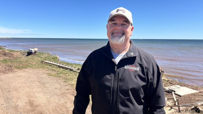 Bernard Savoie est debout devant la mer. Il porte une casquette. Il sourit.