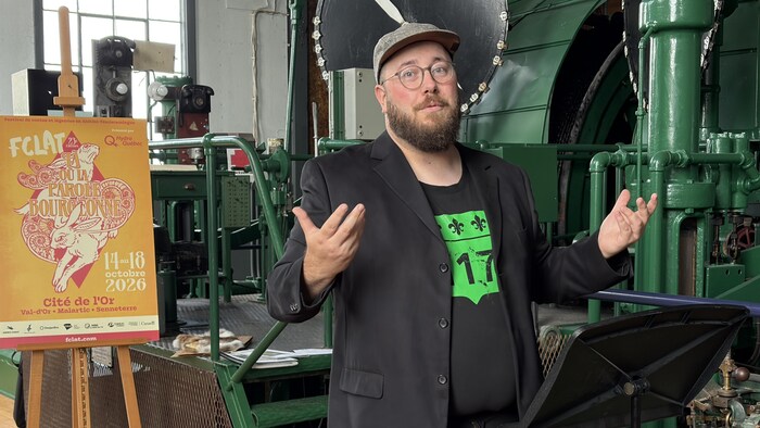Erwann Boulanger parle derrière un lutrin dans la salle du treuil de la Cité de l'Or, devant l'affiche du Festival de contes et légendes.