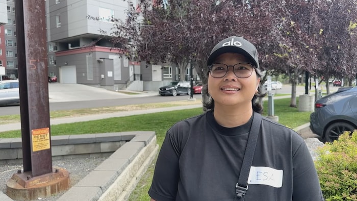 Une femme portant une casquette noire, des lunettes et un chandail noir se tient à l’extérieur, devant un immeuble résidentiel. Une étiquette portant le prénom « ESA » est fixée sur son chandail. On aperçoit derrière elle des arbres, des voitures stationnées et des bâtiments.