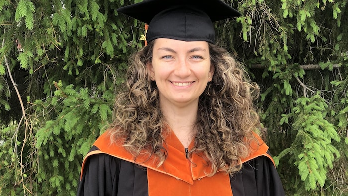 Elsa Dejoie, en uniforme de graduée, sourit à la caméra en posant devant un conifère.