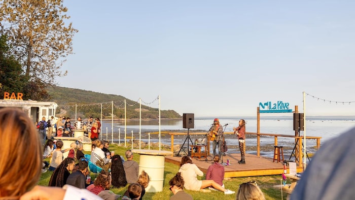 Spectacle de musique en plein air en fin d'après-midi au bord de l'eau à La Rive, Charlevoix. Un duo guitare-violon joue sur scène devant un public assis sur l'herbe, avec le fleuve et des collines en arrière-plan sous un ciel clair.