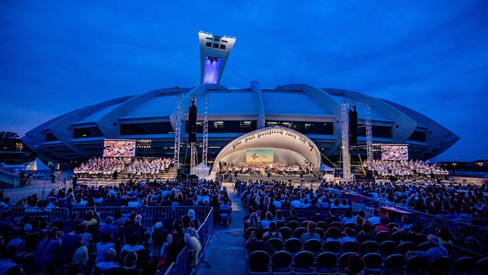 L'Orchestre symphonique de Montréal joue dans un amphithéâtre érigé devant le stade olympique. Des milliers de spectateurs et spectatrices assistent au concert.