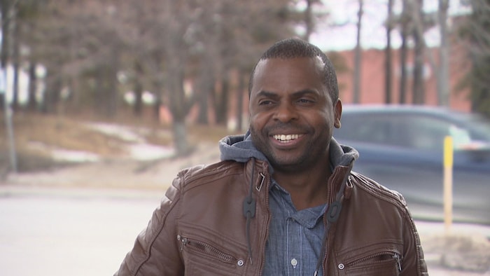 Un homme souriant, un véhicule derrière lui.