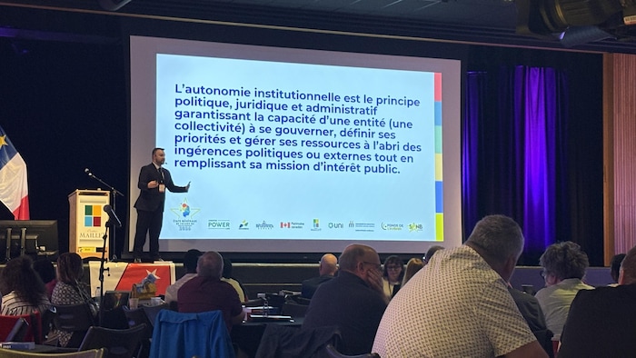 Un homme est debout sur une scène avec un micro et, à côté de lui, une définition du concept d'autonomie institutionnelle est projetée sur écran géant.