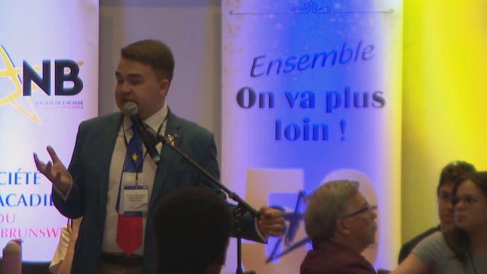 Un homme portant une cravate aux motifs du drapeau acadien est debout et regarde sa main.