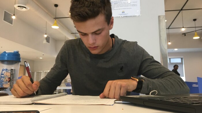 Un jeune attablé rédige des notes non loin de son ordinateur portable.  