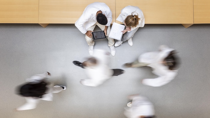 Des personnes en sarrau marchent dans un corridor; d'autres sont assises et regardent une tablette et un cahier.