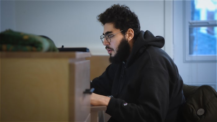 A young man sits at his desk looking at his computer screen.