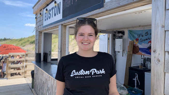 Une femme portant le t-shirt noir du restaurant, devant une cabane en bois, dehors sur la plage.