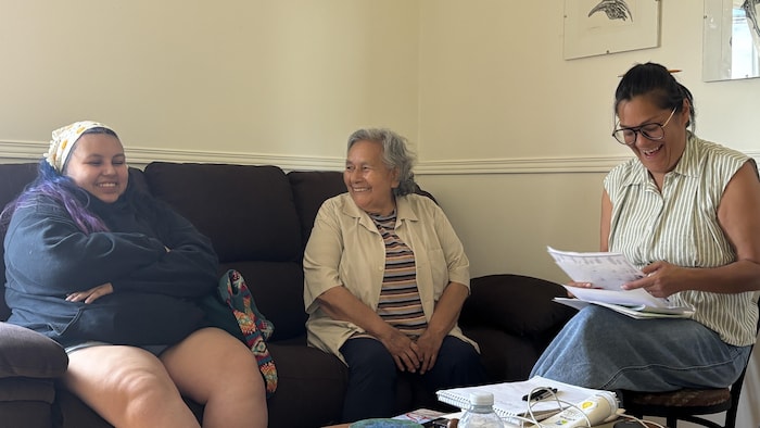 Trois femmes assises côte à côte sur un canapé sourient et rient ensemble dans le salon de la résidence de création.