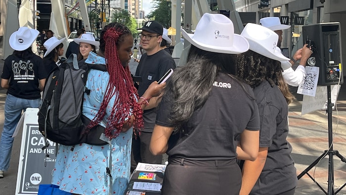 Une passante prend des informations auprès de deux femmes portant un chapeau blanc.