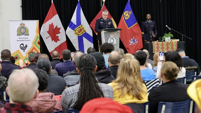 Un gendarme en uniforme est sur une estrade, la tête inclinée sur son discours, pendant qu'une audience l'écoute.