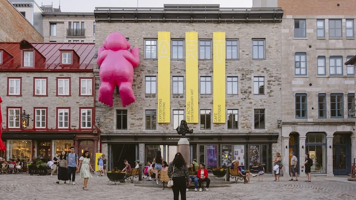 Un imposant personnage gonflable rose escalade la façade en pierre d’un bâtiment historique orné de bannières jaunes.